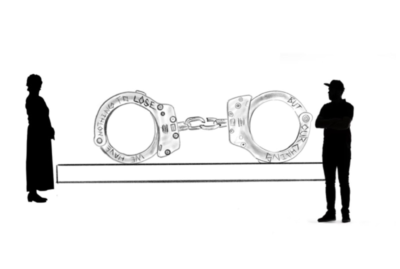 image of two individuals standing on either side of a large scale sculpture of handcuffs