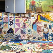 two individuals hol dup a quilt depicting scenes from daily life