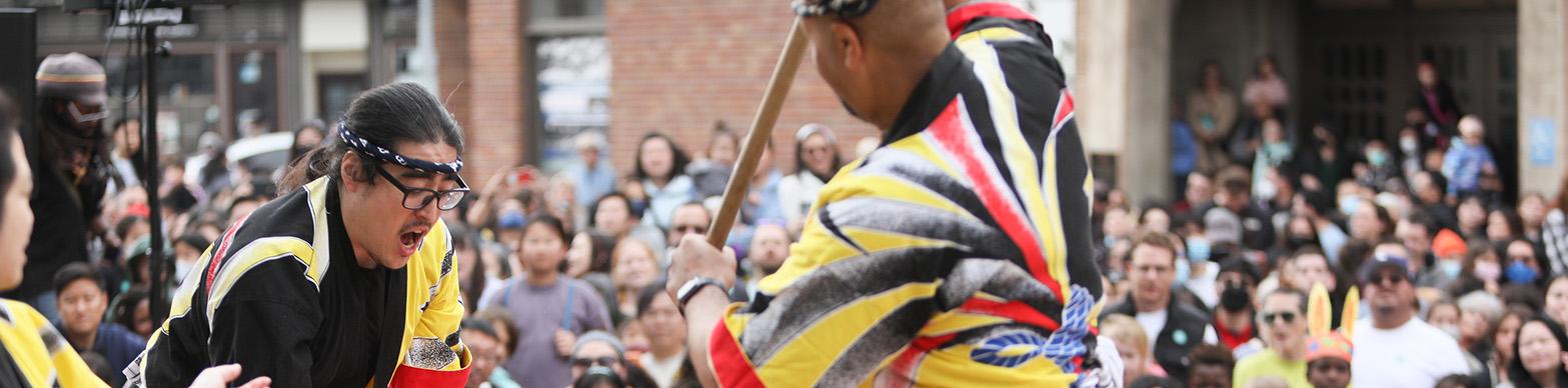 Two japanese men in traditional garb hammering mochi in front of a crowd. 