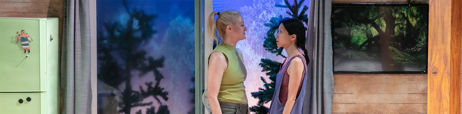 A white woman and an asian woman look into each other's eyes while standing in the door of a cabin