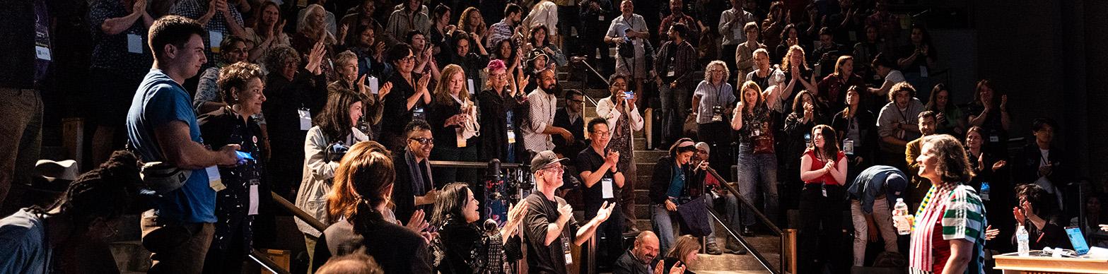 An older woman speaks to a crowded auditorium