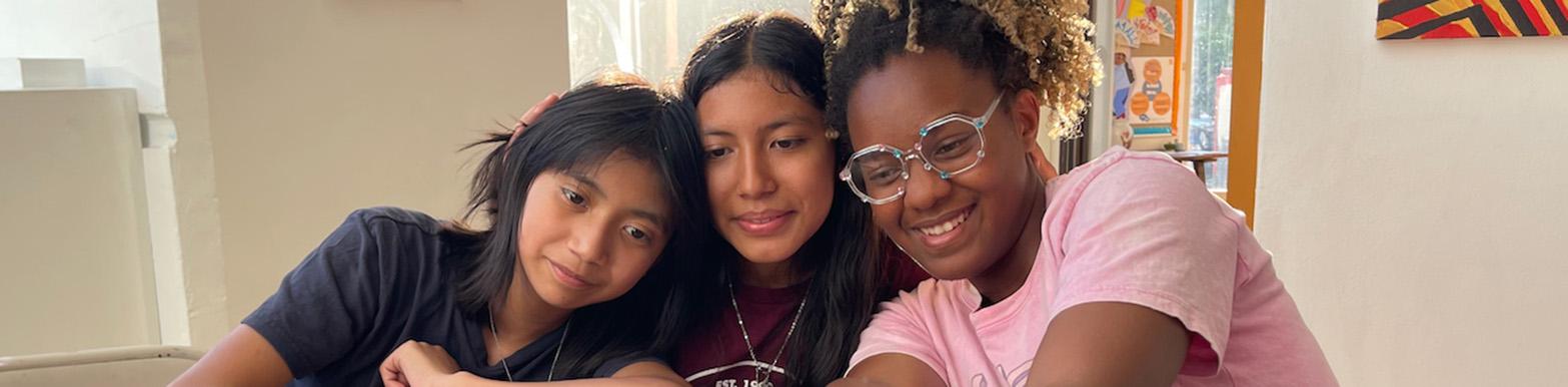 Three young women of multiple races smile and look at a camera they have placed on the table