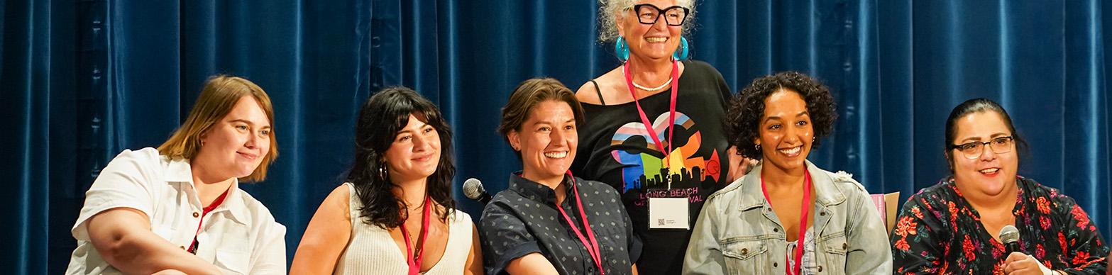 Six women of different ages and races smile for the camera while sitting onstage