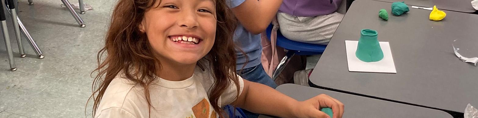 A young girl smiles at the camera while working on a clay sculpture