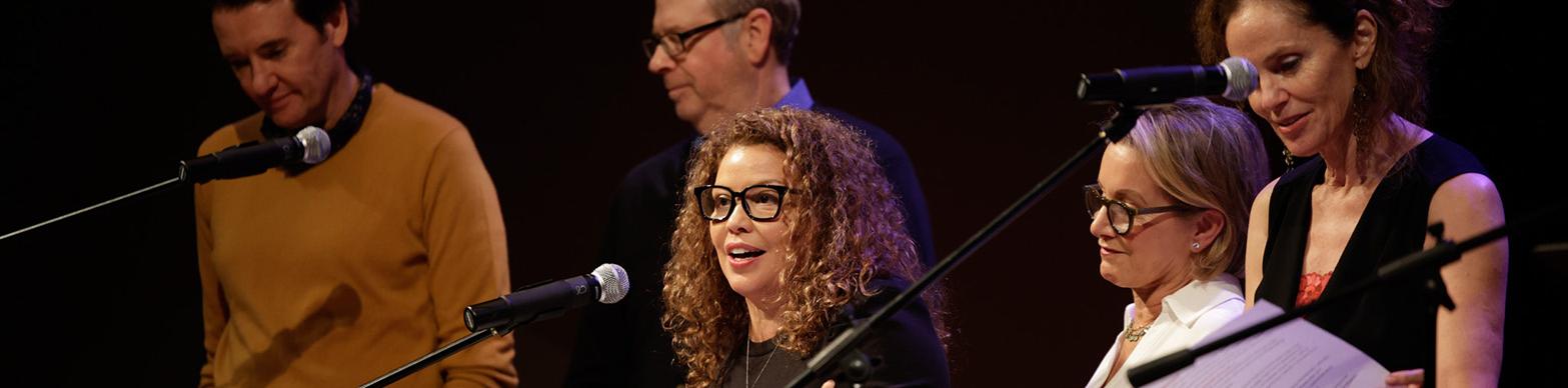Five white individuals read scrips from music stands while standing onstage