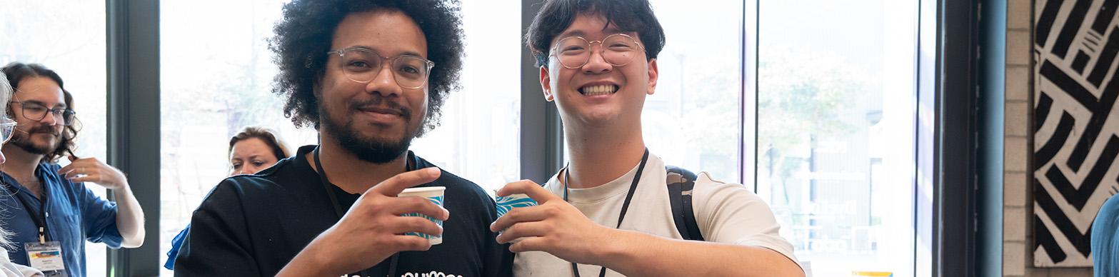 Two young men clink paper cups together as they pose for the camera