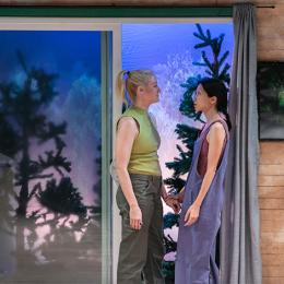 A white woman and an asian woman look into each other's eyes while standing in the door of a cabin