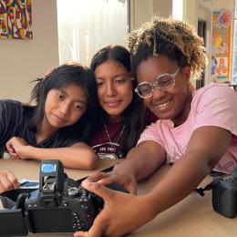 Three young women of multiple races smile and look at a camera they have placed on the table