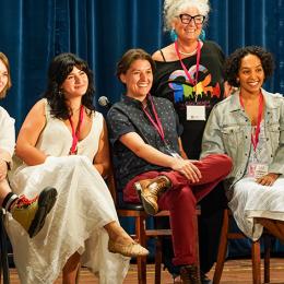 Six women of different ages and races smile for the camera while sitting onstage