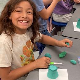 A young girl smiles at the camera while working on a clay sculpture