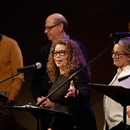 Five white individuals read scrips from music stands while standing onstage